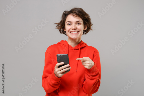 Smiling young brunette woman girl in casual red hoodie posing isolated on grey wall background studio portrait. People lifestyle concept. Mock up copy space. Pointing index finger on mobile phone.