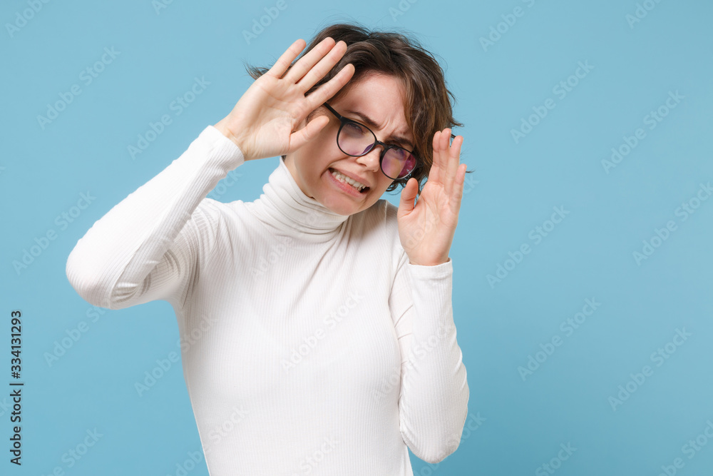 Dissatisfied scared young brunette woman girl in casual white clothes eyeglasses posing isolated on pastel blue background in studio. People lifestyle concept. Mock up copy space. Covering with hands.