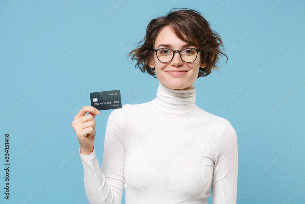 Smiling young brunette woman girl in casual white clothes eyeglasses posing isolated on pastel blue background studio portrait. People lifestyle concept. Mock up copy space. Hold credit bank card.