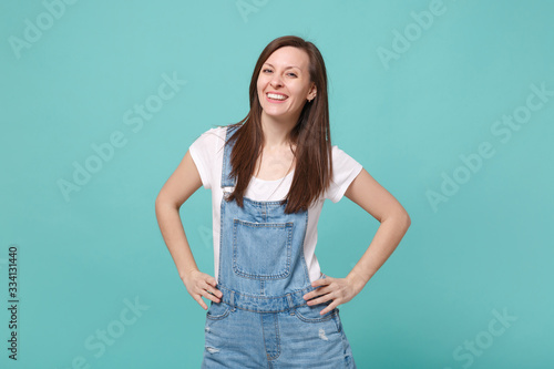 Smiling young brunette woman girl in casual denim clothes posing isolated on blue turquoise background studio portrait. People lifestyle concept. Mock up copy space. Stand with arms akimbo on waist.