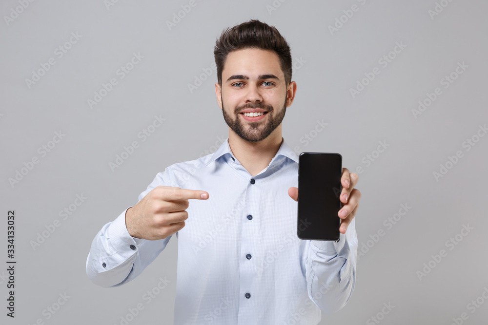 Smiling young unshaven business man in light shirt isolated on grey background. Achievement career wealth business concept. Mock up copy space. Pointing finger on mobile phone with blank empty screen. Smiling young unshaven business man in light shirt isolated on grey background. Achievement career wealth business concept. Mock up copy space. Pointing finger on mobile phone with blank empty screen.
