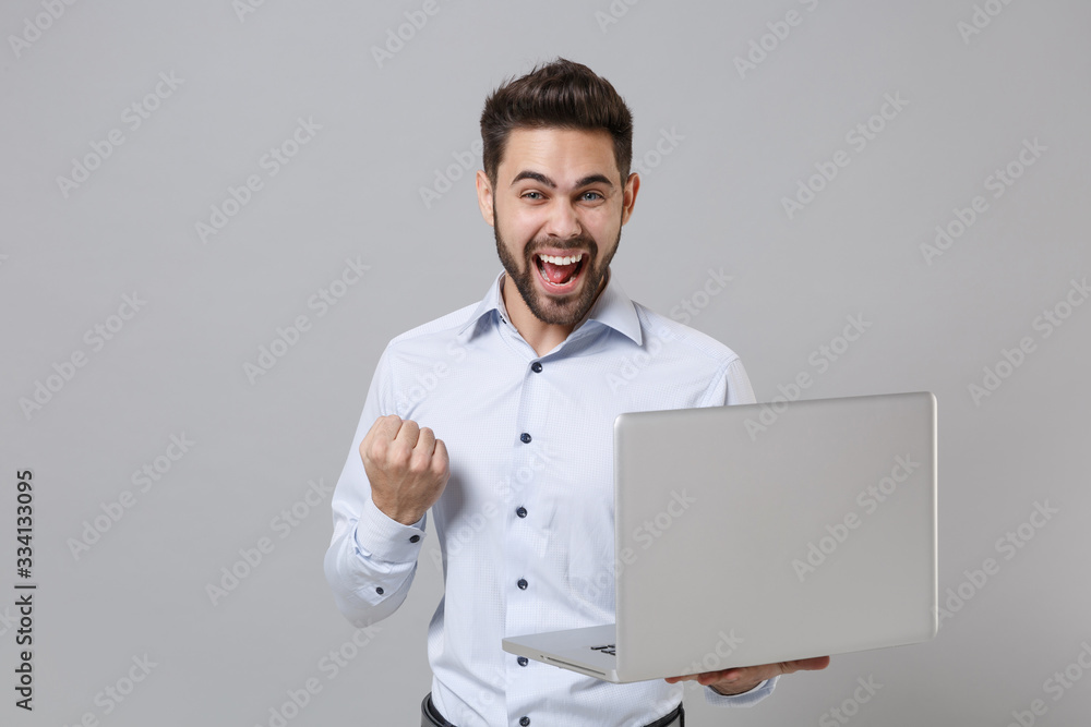 Overjoyed young unshaven business man in light shirt isolated on grey background. Achievement career wealth business concept. Mock up copy space. Working on laptop pc computer, doing winner gesture. Overjoyed young unshaven business man in light shirt isolated on grey background. Achievement career wealth business concept. Mock up copy space. Working on laptop pc computer, doing winner gesture.