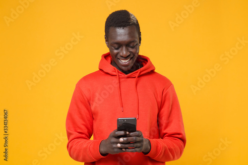 Smiling young african american man guy in red streetwear hoodie posing isolated on yellow background in studio. People lifestyle concept. Mock up copy space. Using mobile phone, typing sms message.