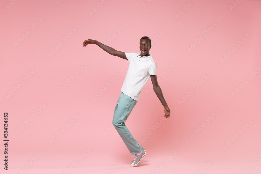 Cheerful young african american man guy in white polo shirt, turquoise trousers posing isolated on pastel pink wall background. People lifestyle concept. Mock up copy space. Standing on toes, dancing. Cheerful young african american man guy in white polo shirt, turquoise trousers posing isolated on pastel pink wall background. People lifestyle concept. Mock up copy space. Standing on toes, dancing.