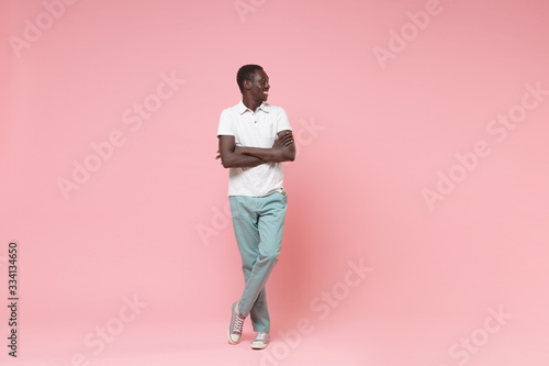 Smiling young african american man in white polo shirt, turquoise trousers posing isolated on pastel pink background. People lifestyle concept. Mock up copy space. Holding hands crossed looking aside.