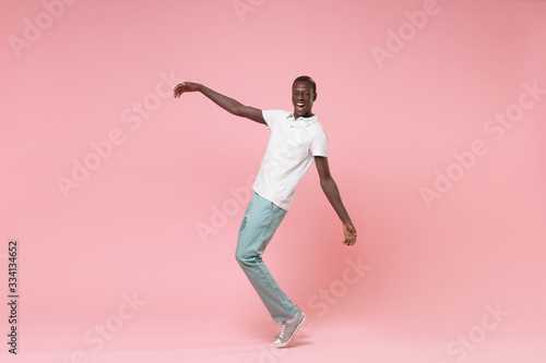 Cheerful young african american man guy in white polo shirt, turquoise trousers posing isolated on pastel pink wall background. People lifestyle concept. Mock up copy space. Standing on toes, dancing.