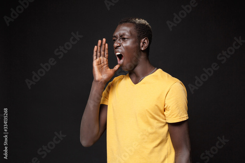 Angry young african american man guy in yellow t-shirt posing isolated on black wall background studio portrait. People lifestyle concept. Mock up copy space. Screaming with hand gesture near mouth.