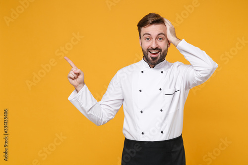 Excited young bearded male chef cook or baker man in white uniform shirt posing isolated on yellow background. Cooking food concept. Mock up copy space. Pointing index finger aside, put hand on head.