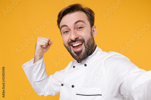Close up of joyful bearded male chef cook or baker man in white uniform isolated on yellow background. Cooking food concept. Mock up copy space. Doing selfie shot on mobile phone doing winner gesture.