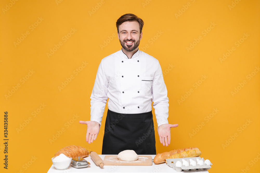 Handsome young bearded male chef cook or baker man in white uniform shirt posing isolated on yellow wall background studio portrait. Cooking food concept. Mock up copy space. Cooking dough at table. Handsome young bearded male chef cook or baker man in white uniform shirt posing isolated on yellow wall background studio portrait. Cooking food concept. Mock up copy space. Cooking dough at table.