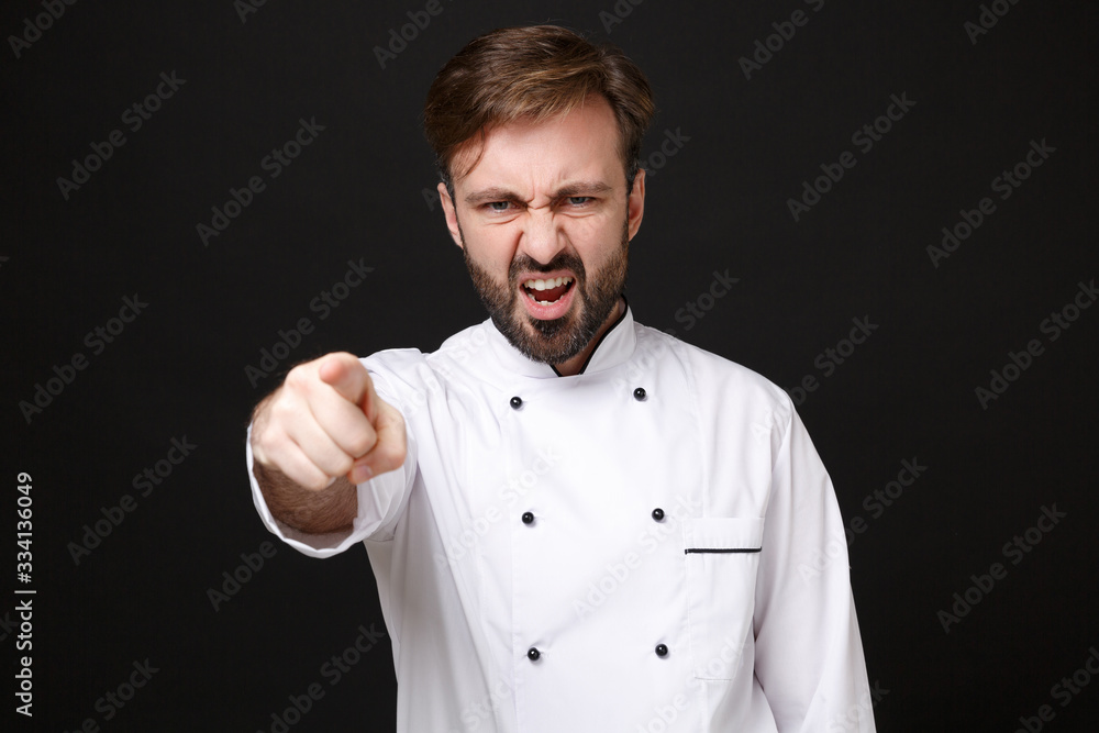 Angry young bearded male chef cook or baker man in white uniform shirt posing isolated on black background. Cooking food concept. Mock up copy space. Point index finger on camera screaming swearing. Angry young bearded male chef cook or baker man in white uniform shirt posing isolated on black background. Cooking food concept. Mock up copy space. Point index finger on camera screaming swearing.