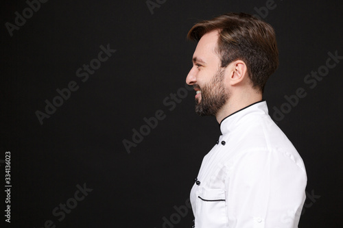 Side view of smiling young bearded male chef cook or baker man in white uniform shirt posing isolated on black wall background studio portrait. Cooking food concept. Mock up copy space. Looking aside.