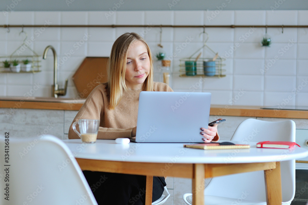 Lovely blonde woman working with her laptop while sitting in the kitchen in her apartment Lovely blonde woman working with her laptop while sitting in the kitchen in her apartment