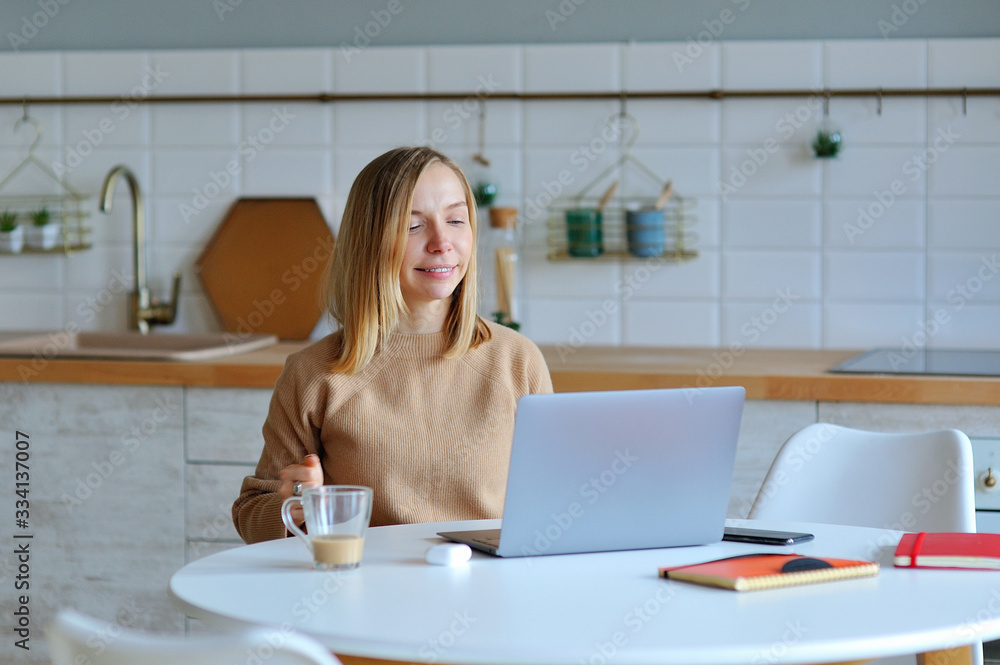 Lovely blonde woman working with her laptop while sitting in the kitchen in her apartment Lovely blonde woman working with her laptop while sitting in the kitchen in her apartment