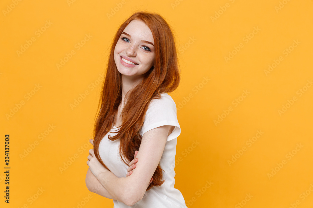 Smiling young redhead woman girl in white blank empty t-shirt posing isolated on yellow wall background studio portrait. People emotions lifestyle concept. Mock up copy space. Holding hands crossed. Smiling young redhead woman girl in white blank empty t-shirt posing isolated on yellow wall background studio portrait. People emotions lifestyle concept. Mock up copy space. Holding hands crossed.