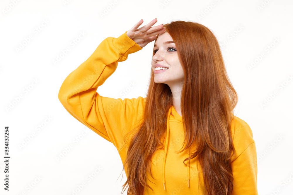 Smiling young redhead woman girl in casual yellow hoodie posing isolated on white background in studio. People lifestyle concept. Mock up copy space. Hold hand at forehead looking far away distance. Smiling young redhead woman girl in casual yellow hoodie posing isolated on white background in studio. People lifestyle concept. Mock up copy space. Hold hand at forehead looking far away distance.