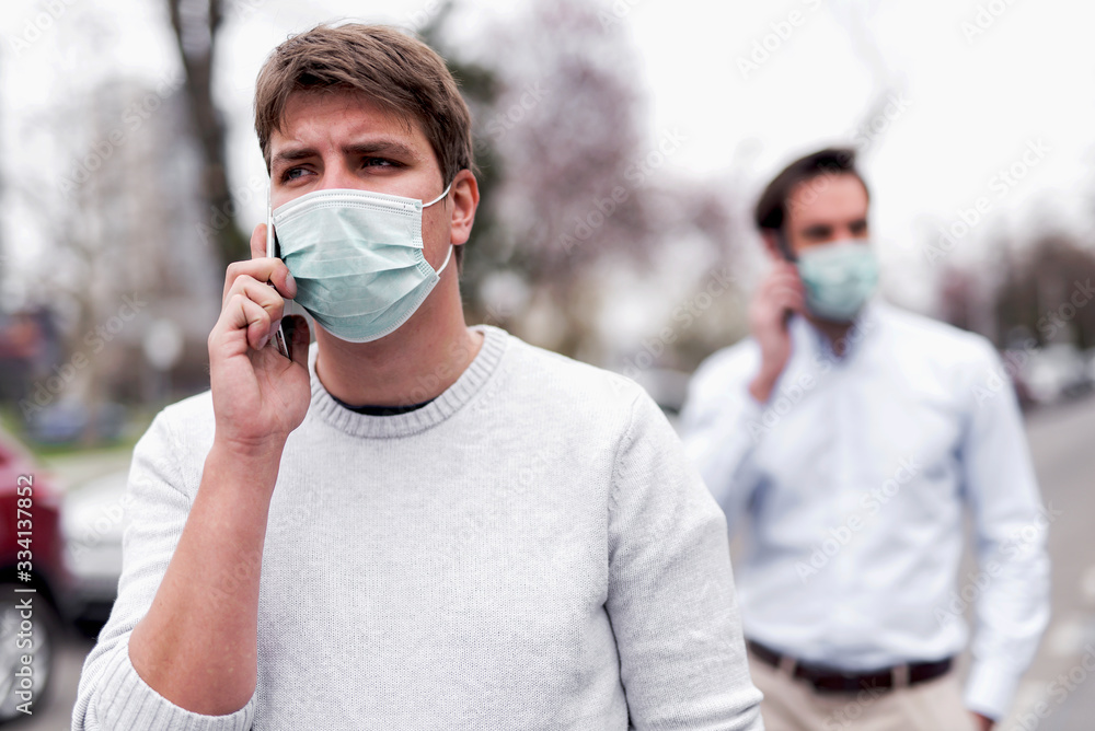 Two businessmen walking outdoor with protective masks and checking them mobiles Two businessmen walking outdoor with protective masks and checking them mobiles