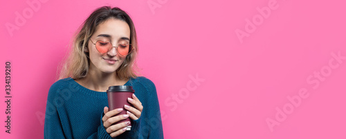 Glamor woman in glasses in a blue sweater with a drink of coffee on a pink background