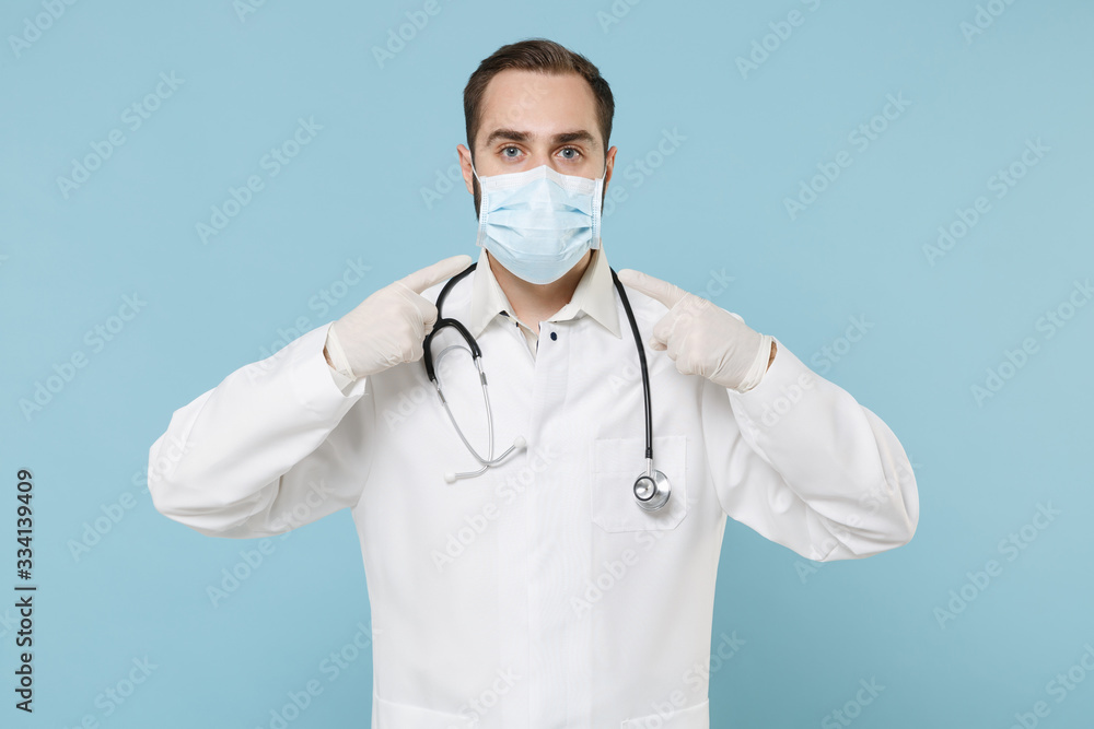 Male doctor man in white medical gown gloves isolated on blue background. Epidemic pandemic spreading coronavirus 2019-ncov sars covid-19 flu virus concept. Point index fingers on sterile face mask. Male doctor man in white medical gown gloves isolated on blue background. Epidemic pandemic spreading coronavirus 2019-ncov sars covid-19 flu virus concept. Point index fingers on sterile face mask.