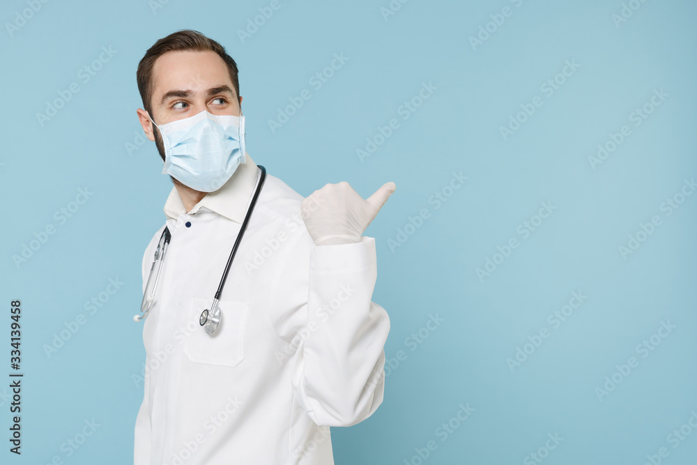 Side view of male doctor man in white medical gown sterile face mask gloves isolated on blue background. Epidemic pandemic coronavirus 2019-ncov sars covid-19 flu virus concept. Pointing thumb aside. Side view of male doctor man in white medical gown sterile face mask gloves isolated on blue background. Epidemic pandemic coronavirus 2019-ncov sars covid-19 flu virus concept. Pointing thumb aside.