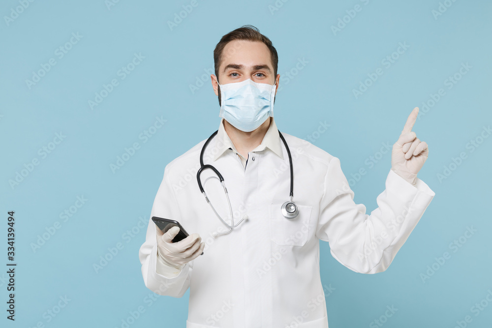 Male on-line doctor man in medical gown sterile face mask gloves isolated on blue background. Epidemic pandemic coronavirus 2019-ncov sars covid-19 flu virus concept. Use mobile phone point finger up.
