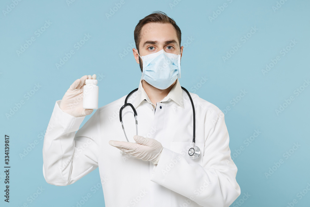 Male doctor man in medical gown face mask gloves isolated on blue background. Epidemic pandemic coronavirus 2019-ncov sars covid-19 flu virus concept. Point hand on medication tablets pills in bottle.