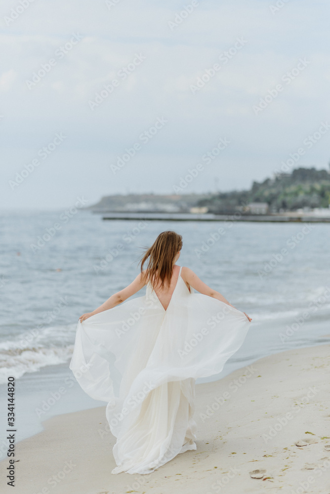 A young beautiful girl in a long milk-colored dress walks along the beach and pier against the background of the sea. A young beautiful girl in a long milk-colored dress walks along the beach and pier against the background of the sea.
