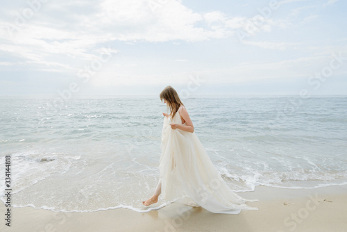 A young beautiful girl in a long milk-colored dress walks along the beach and pier against the background of the sea. © wolfhound911