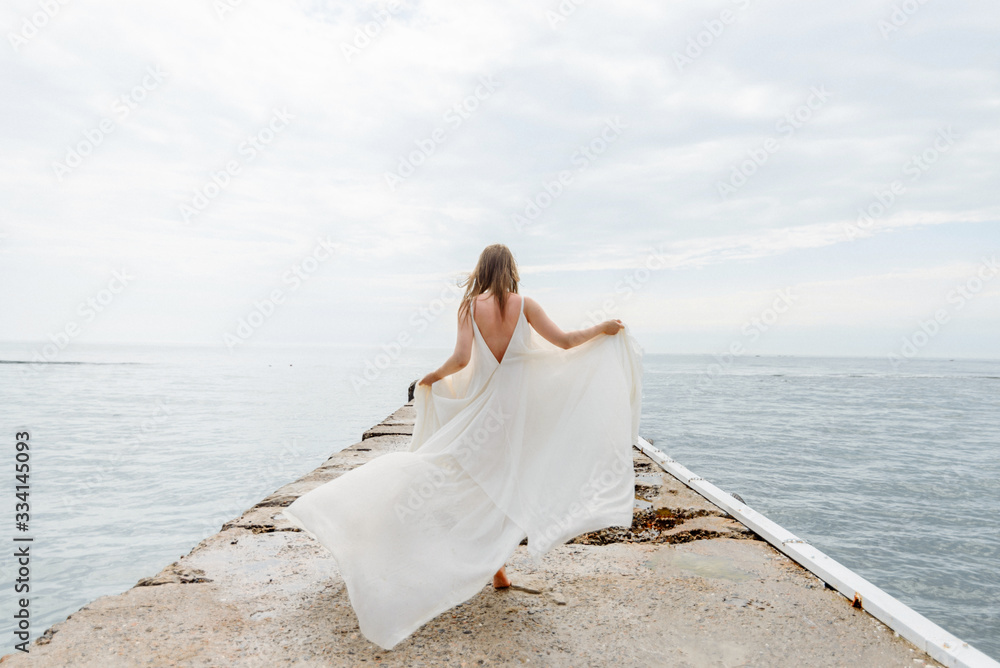 A young beautiful girl in a long milk-colored dress walks along the beach and pier against the background of the sea.