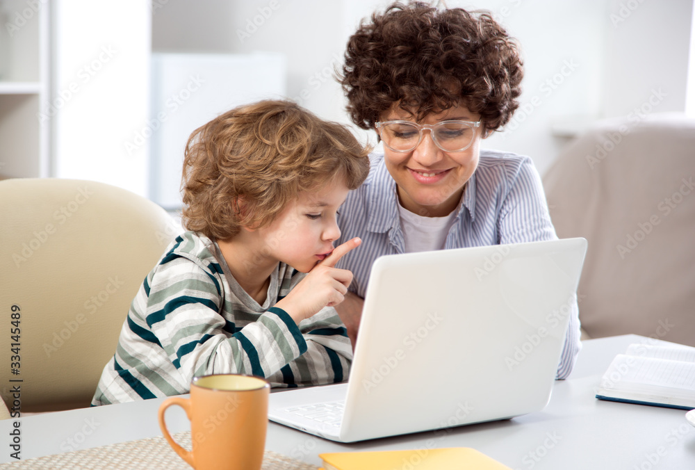 Business woman and her cute little son are using a laptop and smiling while sitting in home office Business woman and her cute little son are using a laptop and smiling while sitting in home office
