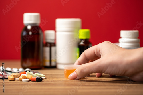 Closeup view photography of female hand taking portion of orange syrup medicine. Many different blurry medical bottles in background.
