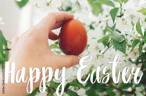 Happy Easter text. Easter greetings lettering. Hand holding modern Easter egg on background of blooming spring cherry flowers. Stylish red egg. Greeting card