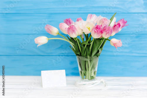 Fototapeta Naklejka Na Ścianę i Meble -  Tulips flowers in glass vase with gift card over blue wooden background for March 8, Women's Day, Birthday, Valentine's Day or Mother's day
