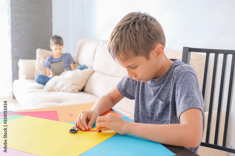 Education, online learning, entertiment at home. Boy making paper craft card with colored paper. Brother using tablet computer for online lesson at home Education, online learning, entertiment at home. Boy making paper craft card with colored paper. Brother using tablet computer for online lesson at home