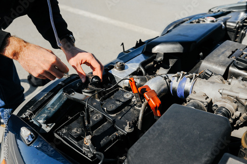 the hand connects the tongs red terminal to the battery of a car with an open hood and tuned parts of the car