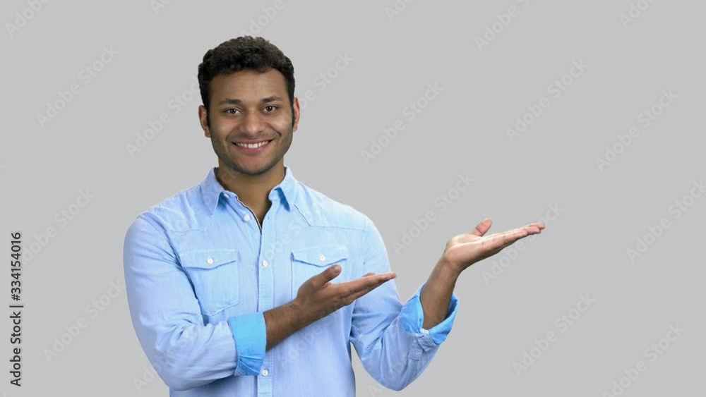 Hindu man shows both palms up. Happy cheerful young guy with dark skin wearing blue shirt. Grey isolated background. Hindu man shows both palms up. Happy cheerful young guy with dark skin wearing blue shirt. Grey isolated background.