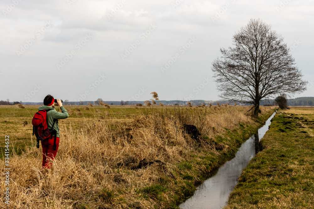 Obraz premium Female tourist person birdwatching in Grady Polewne Natura 2000 area. Woman in red using binoculars in grassland next to channel. Poland, Europe.