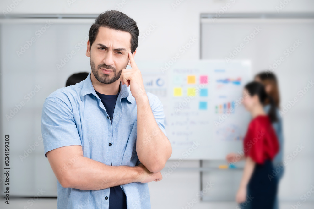 Fototapeta premium Confidence team leader. Young man owner startup business thinking and looking at camera with smile while his colleagues working in the background