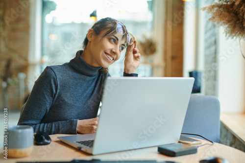 Free Download Attractive pleased woman sitting at the desk with laptop looking away and smiling while working in coffee bar or coworking space. Concept remote work, freelance, using laptop computer or net-book. - GFXLoad