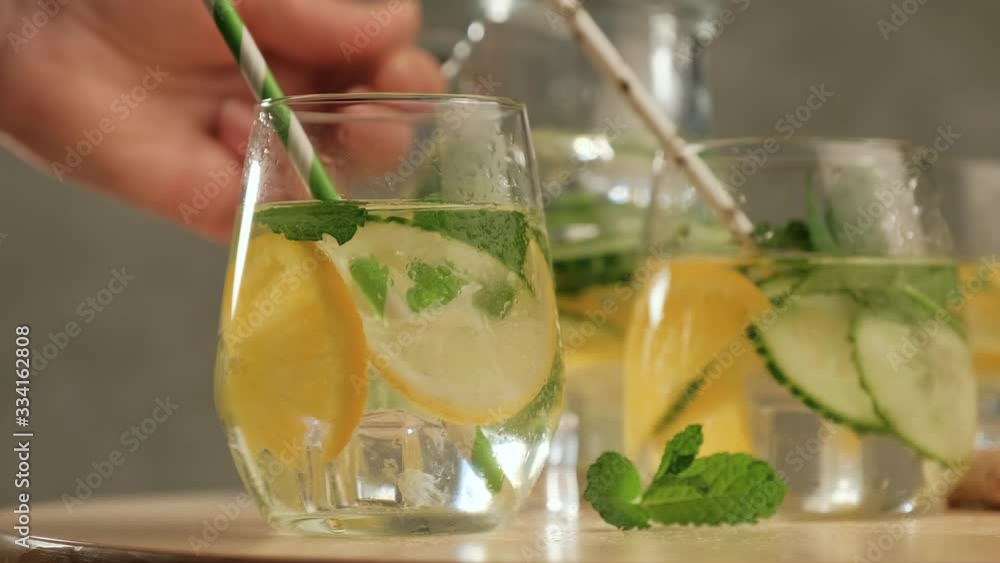 Human hand puts jug of lemonade behind glasses with summer healthy cocktails of citrus infused water with lime lemon orange, ice and mint, diet detox beverages Human hand puts jug of lemonade behind glasses with summer healthy cocktails of citrus infused water with lime lemon orange, ice and mint, diet detox beverages