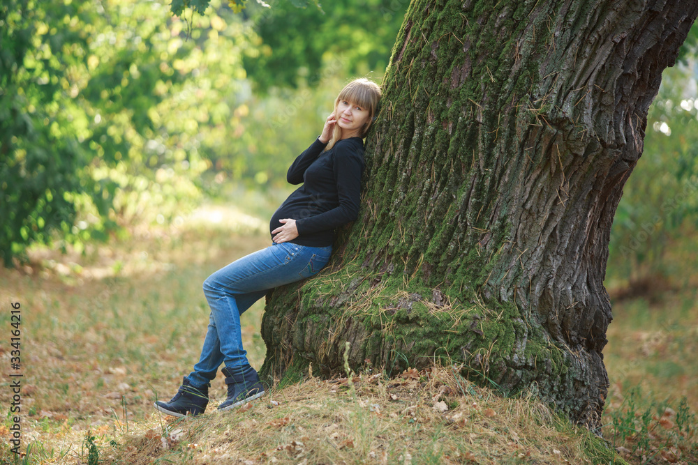 portrait a smiling pregnant woman walking in jeans in the park near the tree. beautiful blonde pregnant woman strokes and touches the belly outdoors in sunny day. happy 9 months of Healthy Pregnancy portrait a smiling pregnant woman walking in jeans in the park near the tree. beautiful blonde pregnant woman strokes and touches the belly outdoors in sunny day. happy 9 months of Healthy Pregnancy