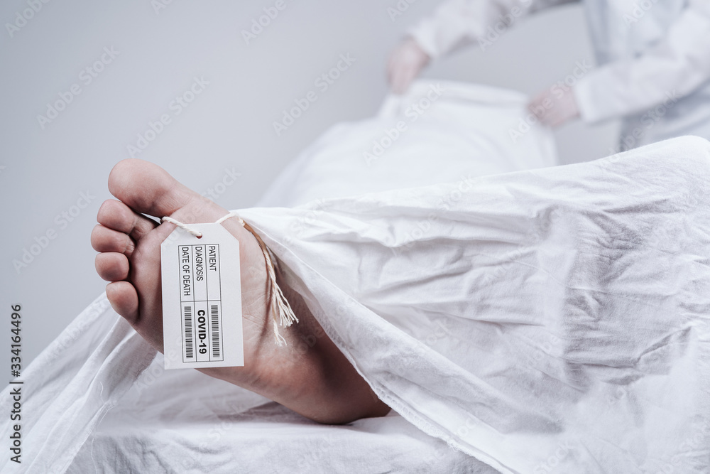 Feet of a dead body, with an identification tag. Deceased's data on sign attached to a big toe. Covered with a white sheet. Virus victim. Feet of a dead body, with an identification tag. Deceased's data on sign attached to a big toe. Covered with a white sheet. Virus victim.
