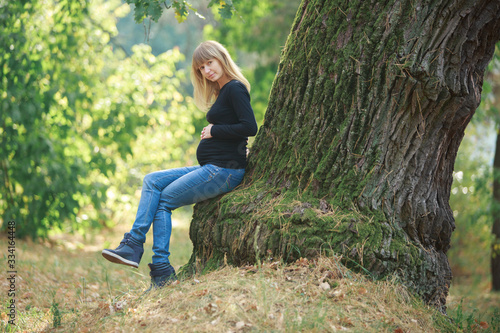 portrait a smiling pregnant woman walking in jeans in the park near the tree. beautiful blonde pregnant woman strokes and touches the belly outdoors in sunny day. happy 9 months of Healthy Pregnancy