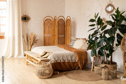 Modern home interior design. White and beige blankets on comfortable bed in boho bedroom interior Textured walls, wooden floor, sunny shades, natural materials, straw screen