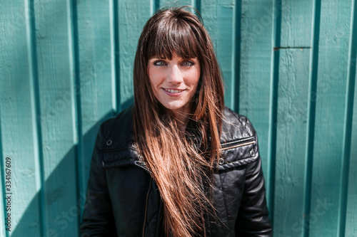 Portrait of a young woman in black coat leaning against a green wooden wall on a sunny afternoon.