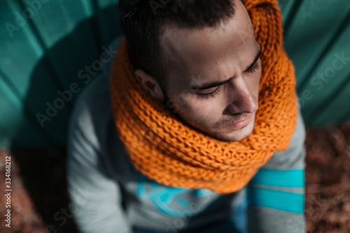 Portrait of a young man sitting on the floor, wearing an orange scarf and leaning against a green wooden wall on a sunny afternoon.