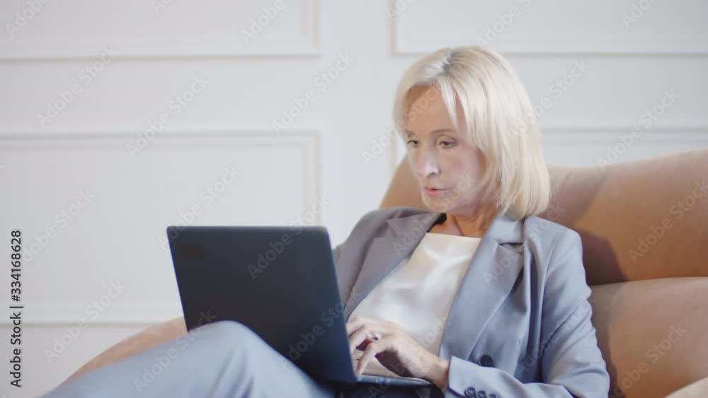 Medium shot of Caucasian blond-haired businesswoman wearing elegant blue pantsuit sitting on comfortable armchair, looking in book and typing on laptop keyboard Medium shot of Caucasian blond-haired businesswoman wearing elegant blue pantsuit sitting on comfortable armchair, looking in book and typing on laptop keyboard