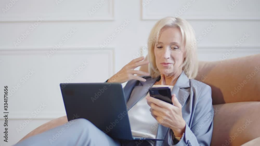 Medium shot of pretty Caucasian businesswoman wearing elegant office clothes sitting on comfortable armchair at home. Woman using laptop and having video conversation using phone Medium shot of pretty Caucasian businesswoman wearing elegant office clothes sitting on comfortable armchair at home. Woman using laptop and having video conversation using phone