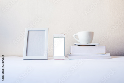 White frame mock-up. Blank frame and mock up screen smartphone with white cup books by the white wall. Mock up frame concept. Minimalism concept. White concept