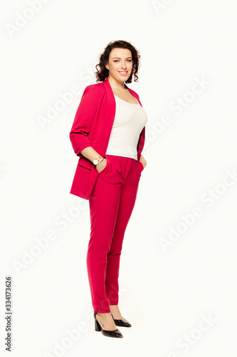 Full length of smiling businesswoman in red suit posing while stands holding hands in pockets and looking at camera isolated over white background.